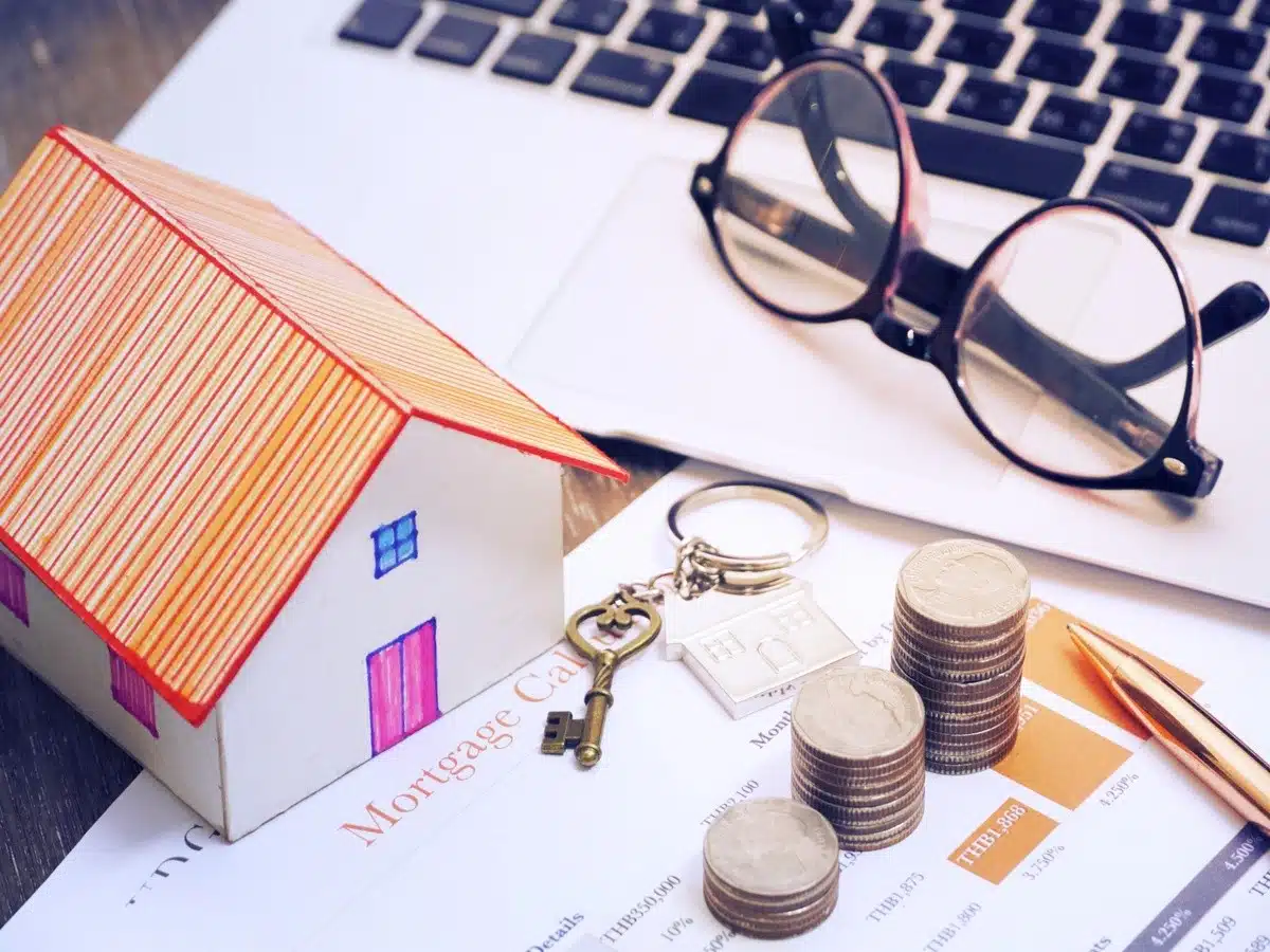 A small paper house, stacked coins, house keychain, glasses, and mortgage paperwork placed on a desk beside a laptop.