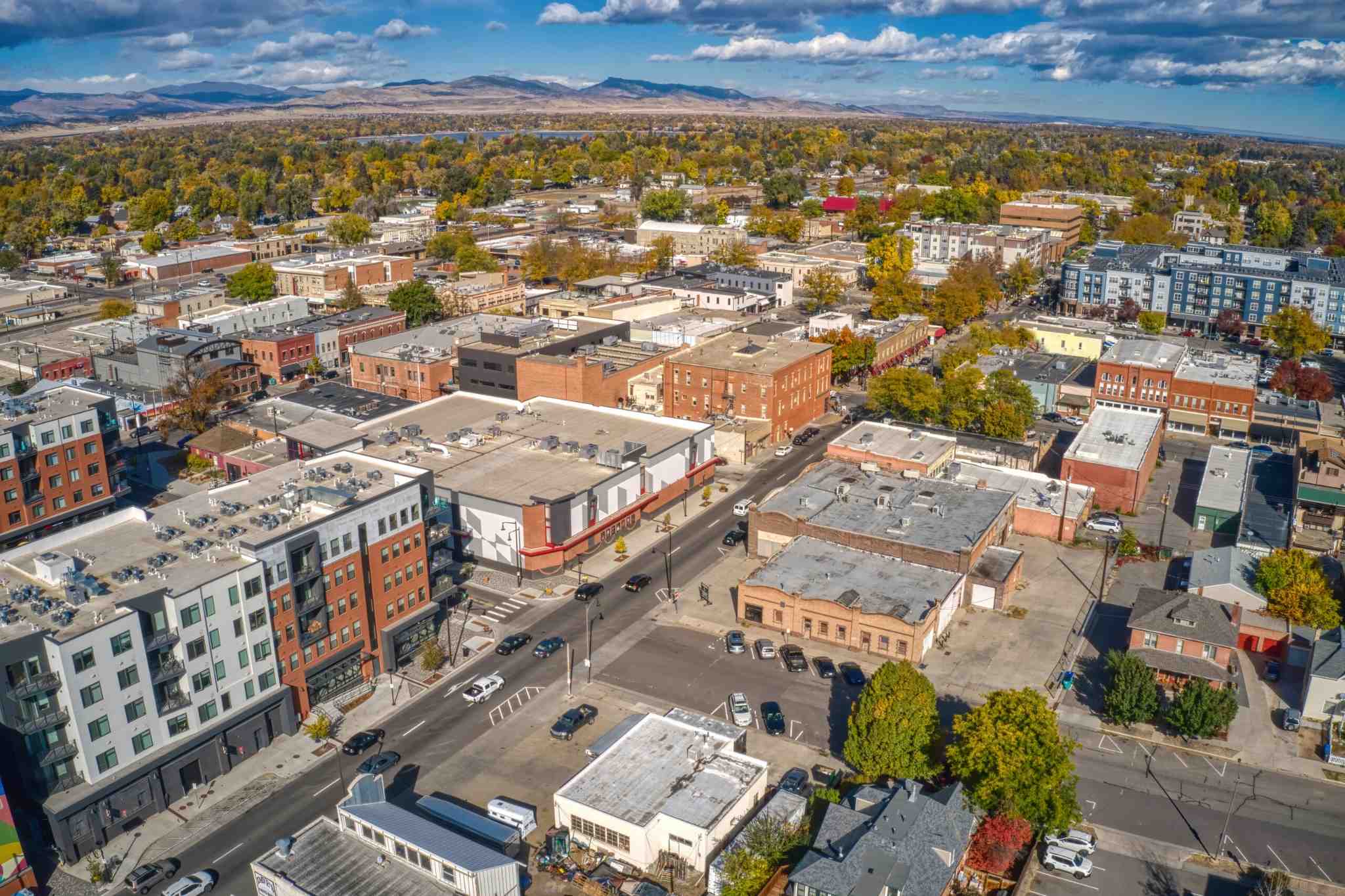 Aerial view of a mid sized Aurora Colorado city with a mix of historic brick buildings, newer apartments, tree lined streets, and distant mountains under a partly cloudy sky.
